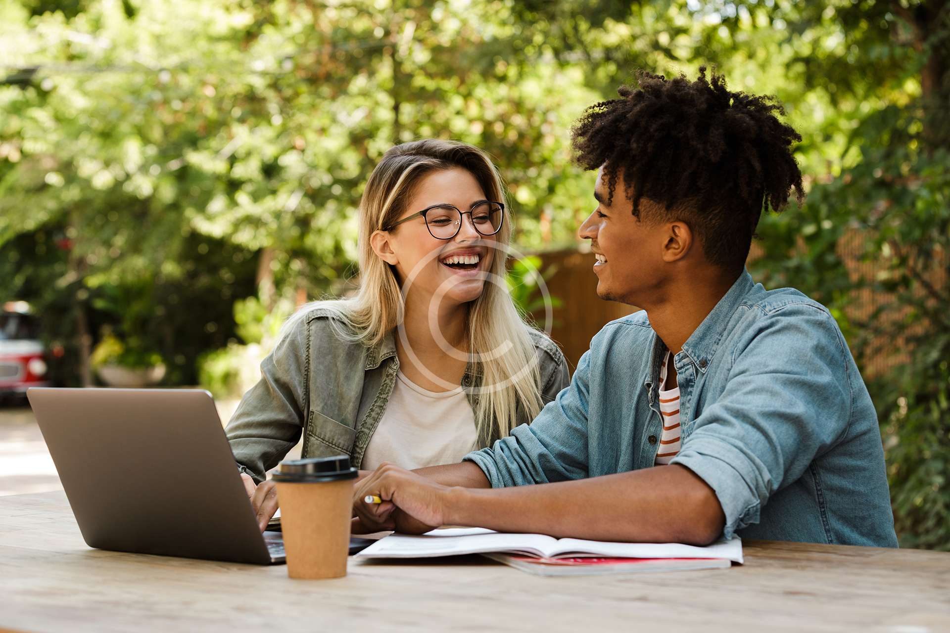 Positive young multiethnic couple spending time together at the park, studying while sitting at the table with laptop